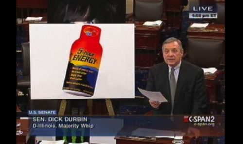 photo of dick durbin speaking on the senate floor, standing next to a huge poster of a 5 hour energy bottle 
