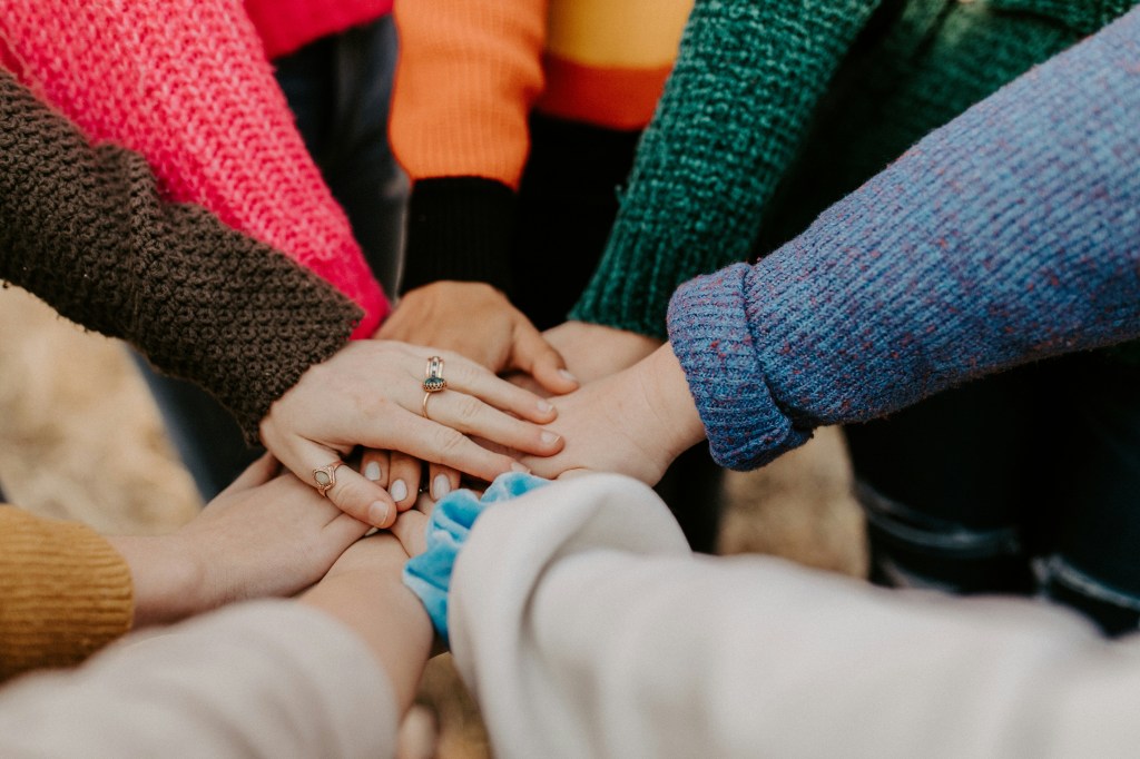 photo of people putting their hands together, each wearing a different, colorful sweater